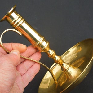 May include: A gold-toned brass candlestick with a curved handle and a round, shallow dish. The candlestick is designed to hold a candle and features a decorative, fluted design.