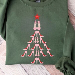 May include: A green sweatshirt with a Christmas tree design made of pink high heels. The tree is topped with a red star.