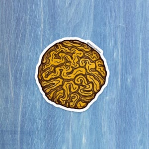 Puede incluir: Una pegatina con un diseño estilizado de cerebro en tonos amarillos y marrones, con un borde blanco. La pegatina está colocada sobre una superficie de madera azul claro. El texto "@dangcuteish" es visible.