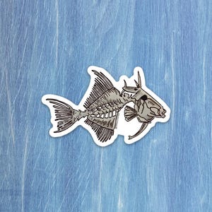 Könnte beinhalten: Ein Aufkleber mit einer detaillierten Illustration eines Fischskeletts in Grau- und Schwarztönen. Das Fischskelett ist mit komplizierten Details dargestellt, einschließlich Wirbelsäule, Rippen und Schädel. Der Aufkleber hat einen weißen Rand und ist vor einem hellblauen Hintergrund platziert.