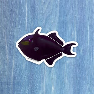 May include: A black fish sticker with a white border. The fish has a rounded body, a dorsal fin, and a tail fin. The fish has a purple eye and a yellow mouth. The text "@DANGCUTEFISH" is printed on the bottom of the sticker.