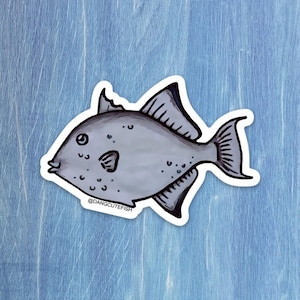 May include: A gray fish sticker with black outlines and details, including fins, a tail, and small spots. The sticker has a white border and the text "@DANGCUTEFISH" at the bottom. The sticker is on a blue wooden surface.