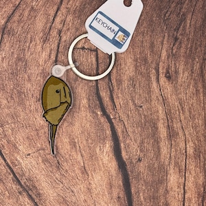 Puede incluir: Un llavero con un diseño de cacerola de herradura. El llavero tiene un accesorio transparente y circular y una etiqueta blanca que dice "KEYCHAIN". La cacerola de herradura es de color marrón y beige, con una ilustración detallada.
