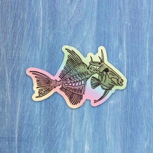 Könnte beinhalten: Ein holografischer Aufkleber mit einer detaillierten Illustration eines Fischskeletts. Das Design umfasst Wirbelsäule, Rippen und Schädel des Fisches mit einem Farbverlauf in Pastellfarben. Der Aufkleber befindet sich auf einer blauen Holzoberfläche.