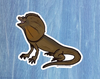 Könnte beinhalten: Ein Aufkleber mit einem Cartoon-Halsbandleguan in Braun- und Beigetönen. Der Leguan hat seinen Kragen ausgebreitet und den Mund geöffnet. Der Aufkleber befindet sich auf einem hellblauen Hintergrund. Der Text "@DANGCUTEFISH" ist sichtbar.