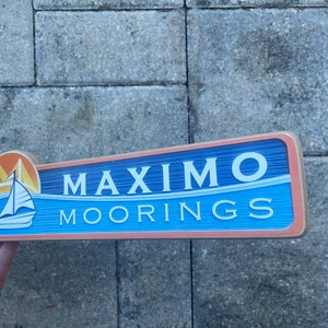 May include: A wooden sign with a blue background and white text that reads "Maximo Moorings". The sign has a small image of a sailboat and a bridge in the upper left corner.