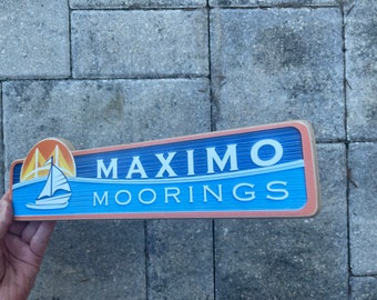 May include: A wooden sign with a blue background and white text that reads "Maximo Moorings". The sign has a small image of a sailboat and a bridge in the top left corner.