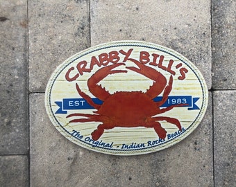May include: A round, oval-shaped sign with a wooden background and blue rope trim. The sign says "Crabby Bill's" in a white, cursive font. A red crab is in the centre of the sign. The sign also says "Est. 1983" and "The Original - Indian Rocks Beach".