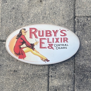 May include: Oval metal sign with the text "Ruby's Elixir & Central Cigars". The sign features a pin-up style illustration of a woman in a red dress holding a cocktail and a cigar. The background is white.
