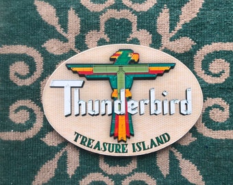 May include: A wooden oval sign with the words "Thunderbird" and "Treasure Island" in white letters. A stylised green, yellow and red bird with outstretched wings is in the centre of the sign.
