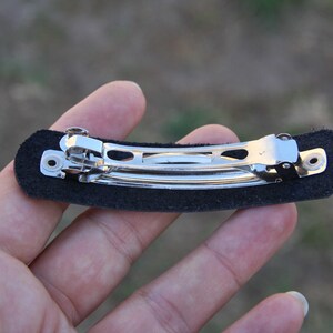 May include: A black barrette hair clip with a silver metal clasp. The clip is attached to a rectangular black felt base. The barrette is designed to hold hair in place.