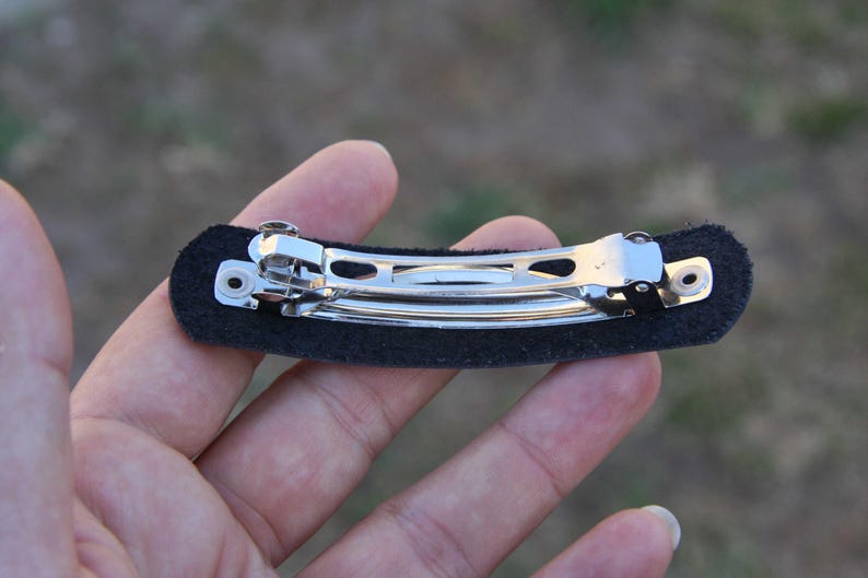 May include: A black barrette hair clip with a silver metal clasp. The clip is attached to a rectangular black felt base. The barrette is designed to hold hair in place.