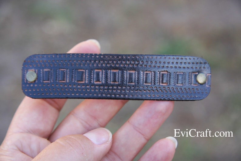 May include: A dark brown leather hair barrette with a rectangular shape. The barrette features a series of embossed rectangular designs and small dot patterns. Two brass-colored rivets secure the barrette.