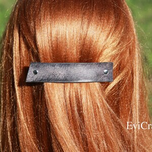 May include: A dark gray leather barrette with two silver rivets, set against a backdrop of auburn hair. The barrette has a rectangular shape with a slightly angled edge. The hair accessory is a stylish addition to any hairstyle.