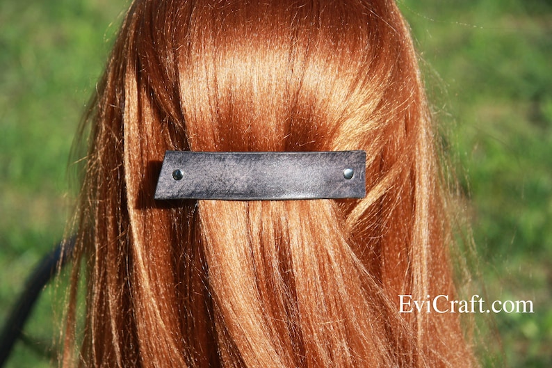May include: A dark gray leather barrette with two silver rivets, set against a backdrop of auburn hair. The barrette has a rectangular shape with a slightly angled edge. The hair accessory is a stylish addition to any hairstyle.