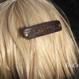 May include: A dark brown leather barrette with a geometric design and brass-colored rivets. The hair accessory is rectangular and is set against a backdrop of blonde hair. The barrette is a hair accessory.
