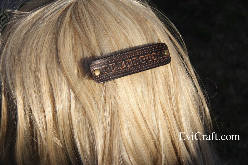 May include: A dark brown leather barrette with a geometric design and brass-colored rivets. The hair accessory is rectangular and is set against a backdrop of blonde hair. The barrette is a hair accessory.