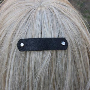 May include: A black leather hair barrette with silver-colored rivets. The barrette is rectangular with rounded edges and is attached to blonde hair. The hair accessory is a simple and elegant design.