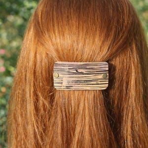 Pode incluir: Uma presilha de cabelo retangular de madeira com um padrão listrado e tachas de cor latão. O acessório de cabelo está preso a cabelos longos e castanhos avermelhados. O fundo é uma vista desfocada de folhagem verde e flores cor-de-rosa.