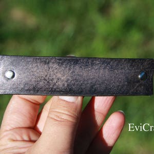 May include: A rectangular, dark gray leather bracelet with a textured surface. The bracelet has a silver rivet on each end. The background is blurred green.