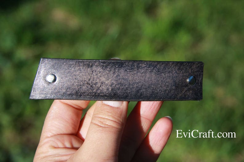 May include: A rectangular, dark gray leather bracelet with a textured surface. The bracelet has a silver rivet on each end. The background is blurred green.