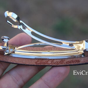 May include: A silver metal barrette hair clip with a brown leather base. The clip is open, revealing the inner workings. The text "EviCraft.com" is visible on the leather. The barrette is designed to hold hair in place.