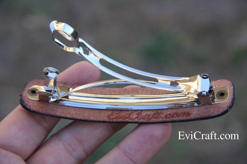 May include: A silver metal barrette hair clip with a brown leather base. The clip is open, revealing the inner workings. The text "EviCraft.com" is visible on the leather. The barrette is designed to hold hair in place.