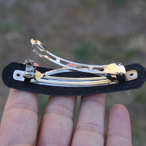 May include: A silver metal hair barrette with a black felt base. The barrette has a hinged clasp and is designed to hold hair in place. The felt base is rectangular and provides a surface for the barrette to attach to the hair.
