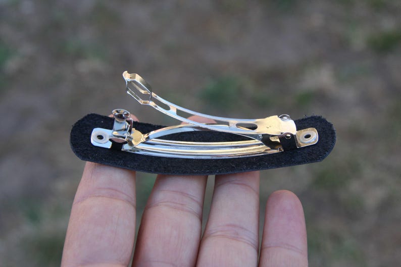 May include: A silver metal hair barrette with a black felt base. The barrette has a hinged clasp and is designed to hold hair in place. The felt base is rectangular and provides a surface for the barrette to attach to the hair.