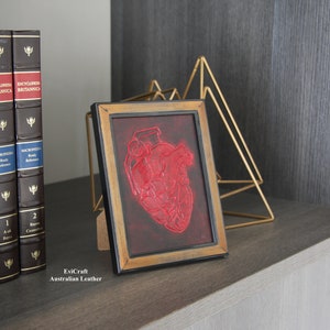 Puede incluir: Una obra de arte enmarcada con un diseño de corazón rojo texturizado sobre un fondo oscuro. El marco tiene un acabado dorado. La obra se exhibe en una estantería con enciclopedias y una escultura geométrica dorada.