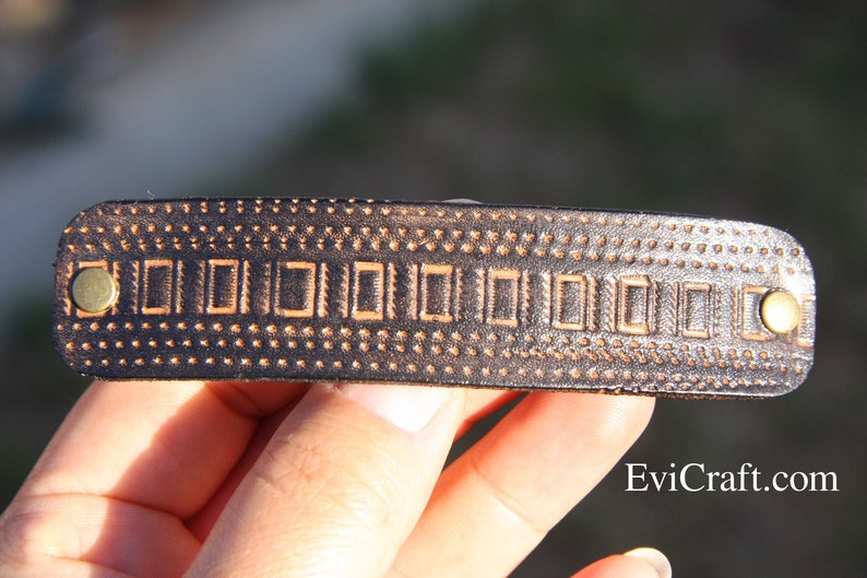May include: A dark brown leather hair barrette with a rectangular design and brass rivets. The barrette features a repeating pattern of small dots and rectangular shapes. The website Evicraft.com is visible.