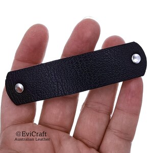 May include: A black leather strap with a textured surface, secured with two silver rivets. The strap is rectangular and appears to be a small accessory or component. The image is a close-up, highlighting the material and hardware.