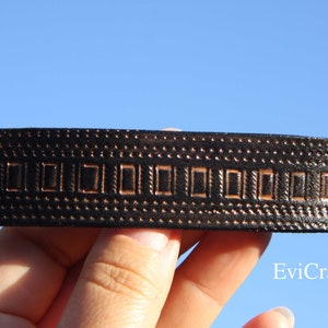 May include: A dark brown leather hair barrette with a rectangular design and brass-colored rivets. The barrette features a repeating pattern of small dots and rectangular shapes. The background is a clear blue sky.