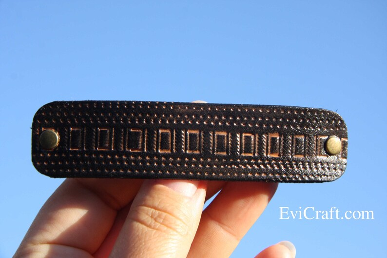 May include: A dark brown leather hair barrette with a rectangular design and brass-colored rivets. The barrette features a repeating pattern of small dots and rectangular shapes. The background is a clear blue sky.