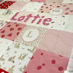 Può includere: Una trapunta patchwork rosa e bianca con una varietà di motivi, tra cui cuori, fiori, fragole e coniglietti. La trapunta è personalizzata con il nome "Lottie" e la data "Nata nel 2021".