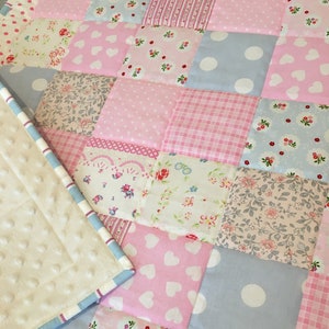 Könnte beinhalten: Ein Patchwork-Quilt mit Quadraten aus rosa, blauen und weißen Stoffen. Die Muster umfassen Punkte, Herzen, Blumendrucke und Streifen. Der Quilt ist mit einem hellblau-weiß gestreiften Rand und einer cremefarbenen, strukturierten Rückseite versehen.