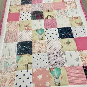 Könnte beinhalten: Ein Patchwork-Quilt mit quadratischen Mustern in Rosa, Weiß und Beige. Die Quadrate zeigen Designs wie Pfeile, Herzen und Blumenmuster. Der Quilt hat einen weißen Rand und eine rosa gepunktete Bordüre.
