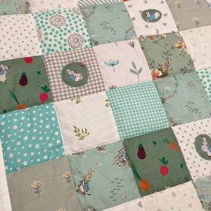 May include: A patchwork quilt with a green and white color scheme. The quilt is made up of squares of different fabrics, including floral prints, polka dots, stars, and stripes. The quilt features a Peter Rabbit theme with images of Peter Rabbit and other characters from the story.