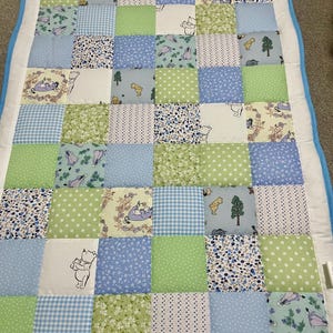 Könnte beinhalten: Ein Patchwork-Quilt mit Quadraten in verschiedenen Mustern und Farben, darunter Hellblau, Grün und Weiß. Einige Quadrate zeigen Winnie-the-Pooh-Illustrationen. Der Quilt hat einen weißen Rand mit hellblauer Einfassung.