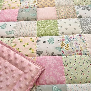 Könnte beinhalten: Ein Patchwork-Quilt mit einer Vielzahl von Pastellstoffen. Der Quilt zeigt Quadrate mit Blumen-, Schmetterlings- und Herzmustern in Rosa-, Grün- und Grautönen. Eine weiche, rosa, strukturierte Decke ist über den Quilt gefaltet.