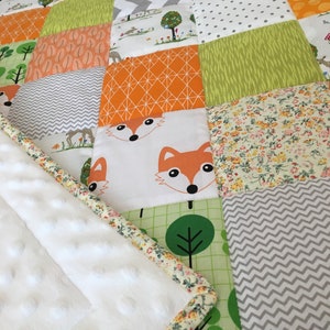 May include: A white baby blanket with a patchwork design featuring orange, green, and gray fabrics. The blanket has a white, plush backing and a floral trim.
