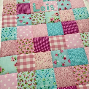 Könnte beinhalten: Eine Patchwork-Decke aus verschiedenen Stoffquadraten in Rosa-, Magenta- und Türkistönen. Die Muster umfassen Blumenprints, Vichy-Karos und Punkte. Der Name Lois ist in Hellblau auf einem der zentralen Quadrate aufgestickt.