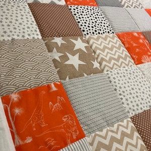 May include: A patchwork quilt with a variety of patterns and colors, including orange, brown, gray, and white. The quilt features a dinosaur pattern, stars, and geometric shapes.