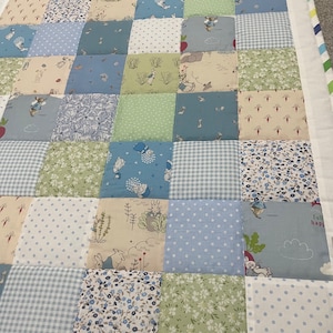 May include: A patchwork quilt featuring a variety of blue, green and white fabrics with a Peter Rabbit theme. The quilt is made up of squares with different patterns, including polka dots, gingham and floral prints. The quilt is edged with a white border.