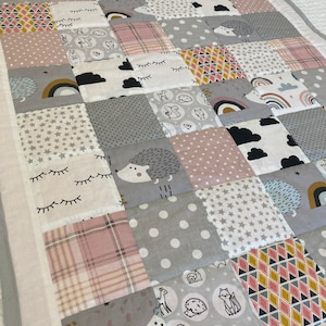 Op de afbeelding: Een patchwork babydeken met verschillende patronen in grijstinten, roze en wit. De deken heeft verschillende ontwerpen, waaronder sterren, wolken, regenbogen en dieren.