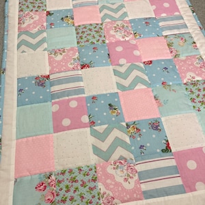 May include: A patchwork quilt with a variety of floral and geometric patterns in shades of pink, blue, and white. The quilt is made of squares and rectangles, and the seams are visible.