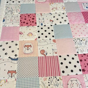 Könnte beinhalten: Eine rosafarbene und blaue Patchwork-Babydecke mit verschiedenen Tier- und geometrischen Mustern. Die Decke zeigt einen Fuchs, eine Katze, einen Pinguin, ein Einhorn, ein Flusspferd, ein Lama, einen Flamingo, einen Regenbogen, Sterne und Punkte.