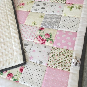 May include: A patchwork baby quilt with a white background and pink, grey, and floral fabric squares. The quilt has a white border and a grey binding. The quilt is made by "The Baby Boutique" and has a tag that says "Handmade with Love".