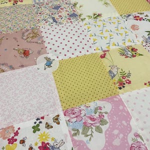 Könnte beinhalten: Eine Patchwork-Decke mit verschiedenen floralen und Hasen-Motiven in Rosa, Gelb und Weiß. Die Decke besteht aus Quadraten aus verschiedenen Stoffen, jedes mit einem einzigartigen Design.