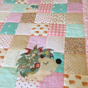 Könnte beinhalten: Eine rosa und grüne Patchwork-Babydecke mit einem Igel-Applikationen. Die Decke hat eine Vielzahl von Mustern, darunter Sterne, Punkte, Streifen und Gingham. Der Name "Amelia" ist auf die Decke gestickt.